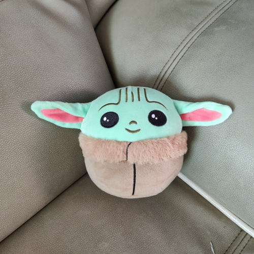 Disney Mandalorian Baby Yoda plush toy with mint green head, pink inner ears, and tan fuzzy body on gray couch